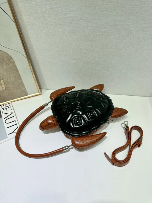 A turtle-shaped shoulder bag with a glossy black embossed shell and brown textured faux-leather legs, flippers, and a matching brown shoulder strap, displayed on a white surface.