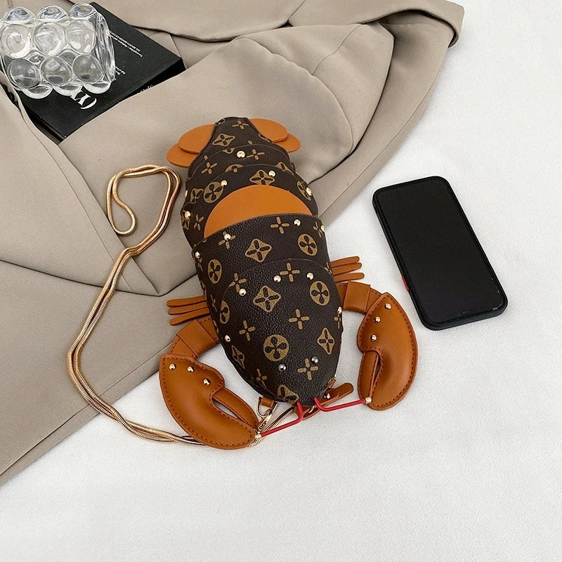 A lobster-shaped crossbody bag with a brown and tan Louis Vuitton-style monogram pattern, styled on a beige blazer next to a smartphone on a white surface.