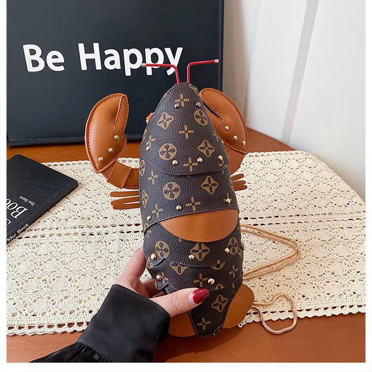 A lobster-shaped handbag with a dark brown and beige monogram pattern, tan leather accents, and a gold chain strap, held by a hand over a lace table runner.