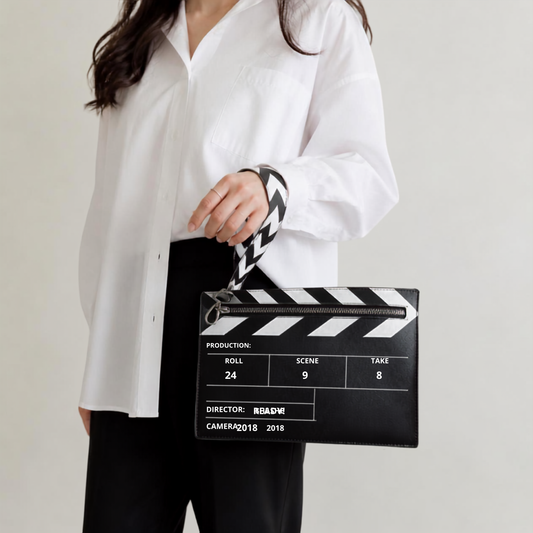 A person wearing a white button-up shirt and black pants holds a black clapperboard-shaped clutch with a black-and-white chevron strap against a plain light background.