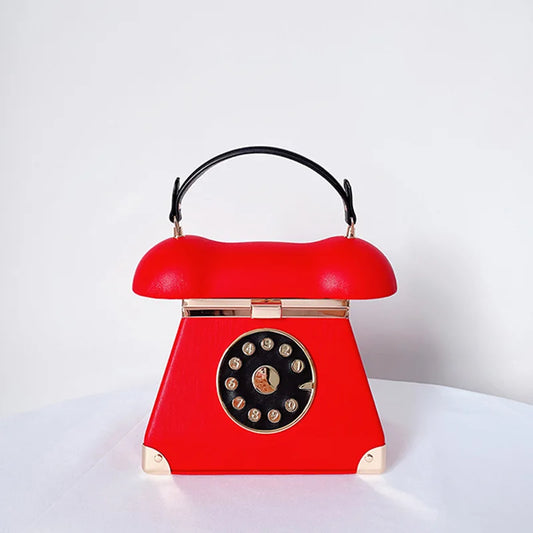 A red vintage rotary telephone handbag with a black handle, gold-tone hardware, and a black circular dial face on a white surface against a white background.