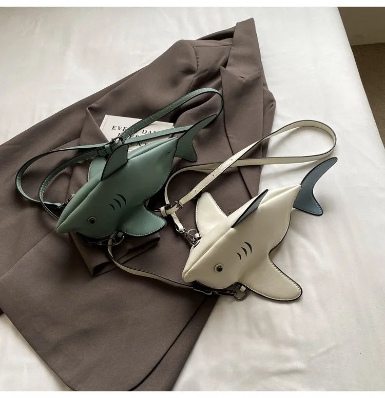 Two shark-shaped crossbody bags, one in sage green and one in cream, displayed on a brown blazer over a white fabric background.