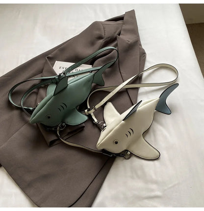 Two shark-shaped crossbody bags, one in sage green and one in cream, displayed on a brown blazer over a white fabric background.