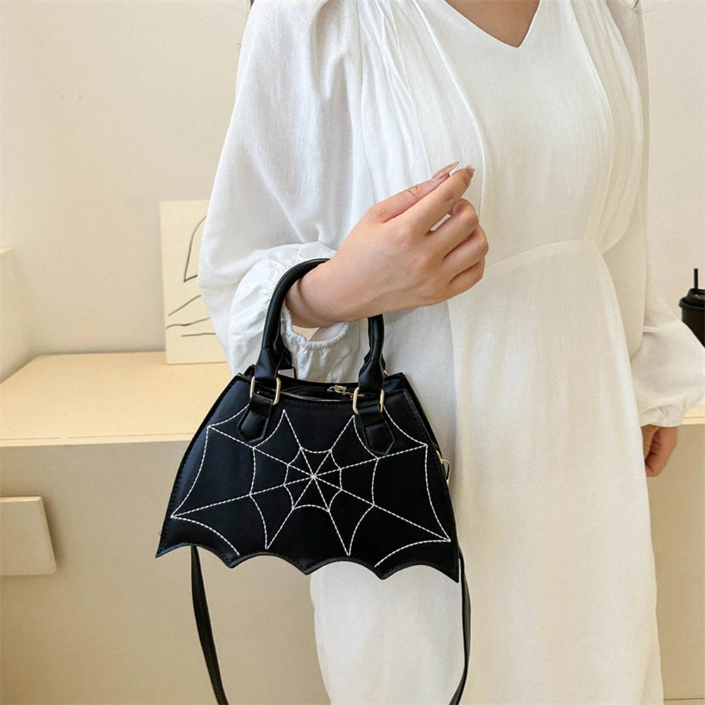Black bat-shaped handbag with spider web design held by a person wearing a white outfit.