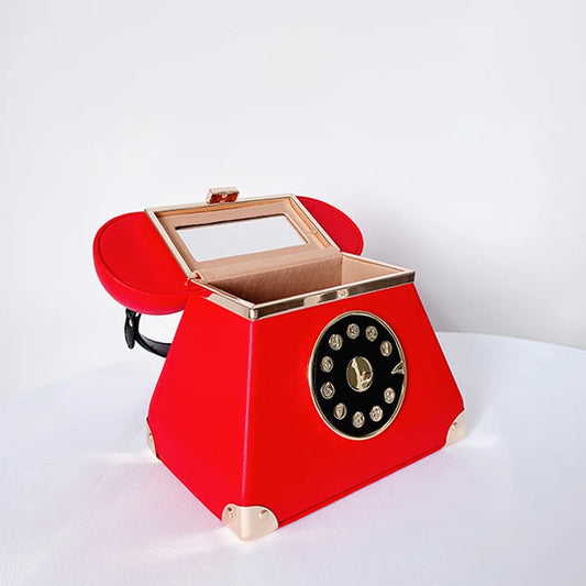 A red novelty jewelry box designed like a vintage rotary telephone, featuring a hinged lid with a clear glass window, gold-tone trim, and a black circular dial face on the front.