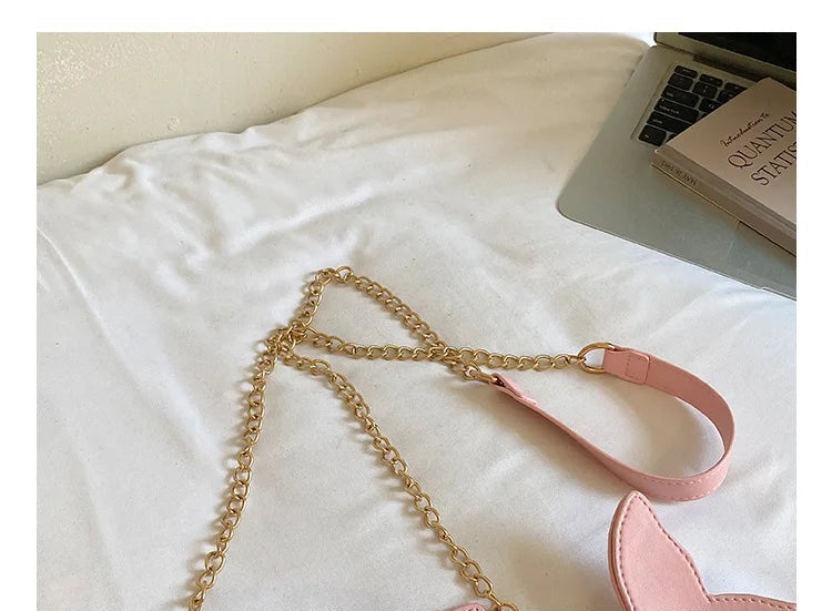 Pink faux-leather shoulder bag with a gold chain strap and a pink shoulder strap, shown on a white fabric surface with a laptop and book in the background.