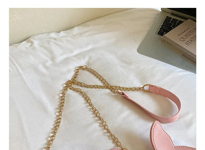 Pink faux-leather shoulder bag with a gold chain strap and a pink shoulder strap, shown on a white fabric surface with a laptop and book in the background.