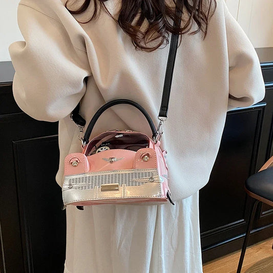 A person wearing a cream-colored sweater and white skirt carries a small pink handbag with a silver metallic front panel and black strap in an indoor setting.
