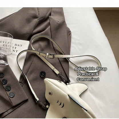 A cream-colored shark-shaped shoulder bag with a silver metal eyelet and black mouth details, styled on top of a brown blazer with a white fabric background. A glass and a book are partially visible on the left.