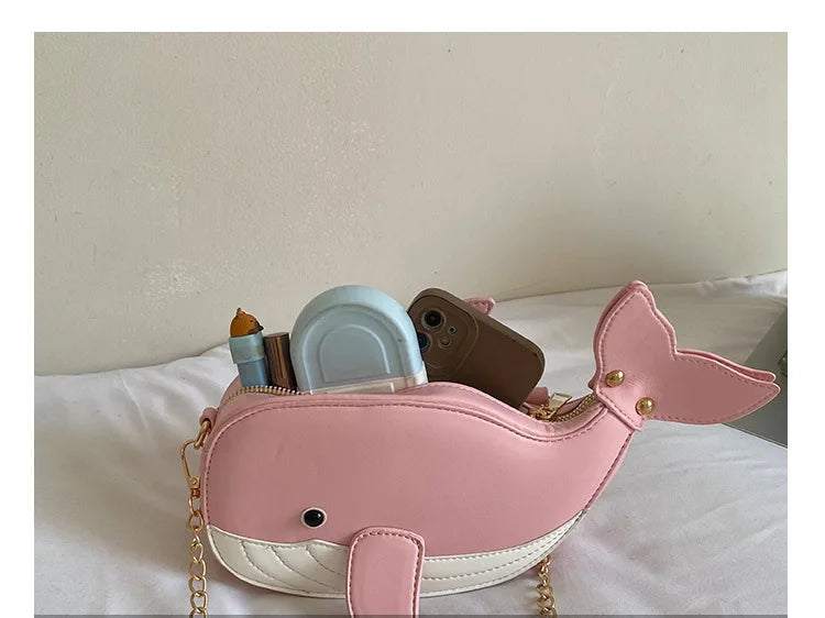 A pink whale-shaped shoulder bag with a white belly, gold-tone chain strap, and small gold hardware, shown resting on a white bed with various items tucked inside its open pouch. Chinese text overlay at the bottom reads '通人税袋' and 'THE DETAILS SHOW'.