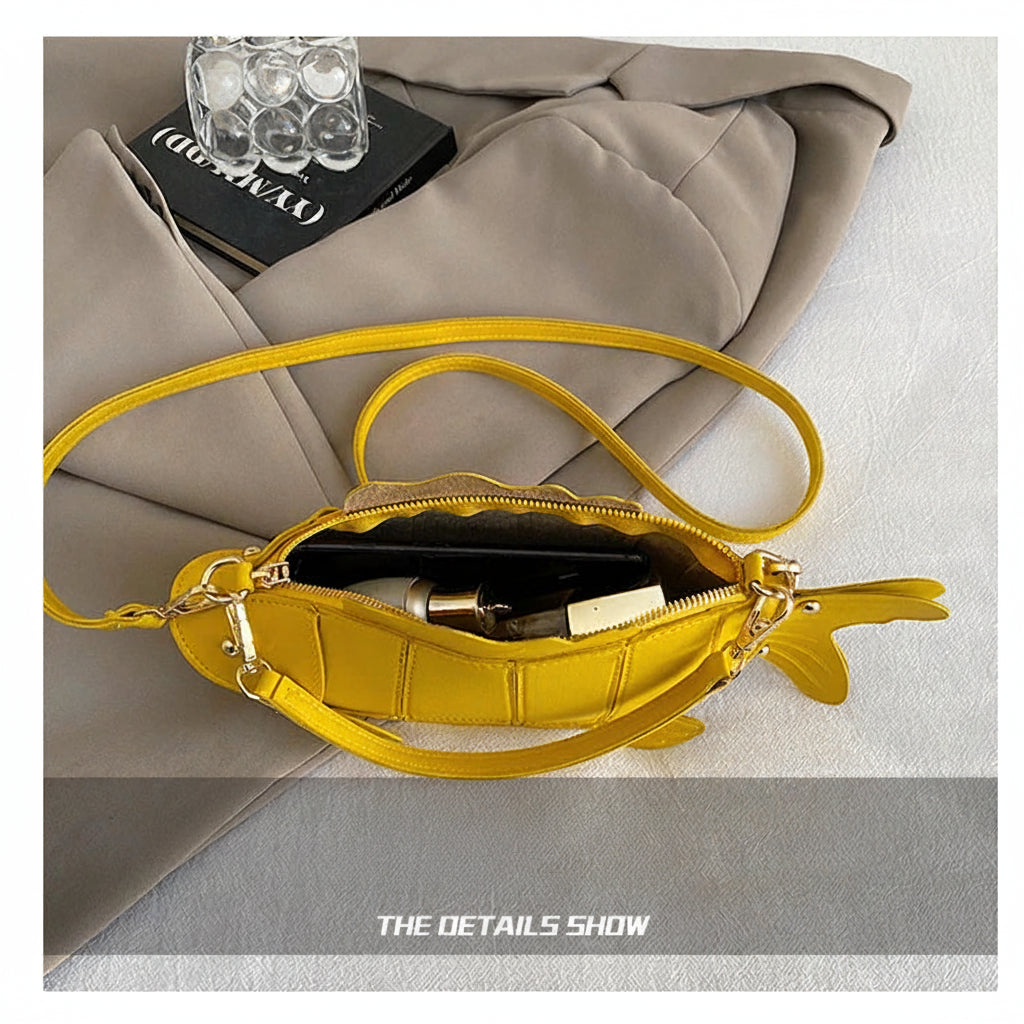 A bright yellow fish-shaped shoulder bag with a zippered body and long strap, shown open with a phone and small accessories inside. The bag is laid on a light gray fabric surface with a folded taupe blazer and a black book in the background.