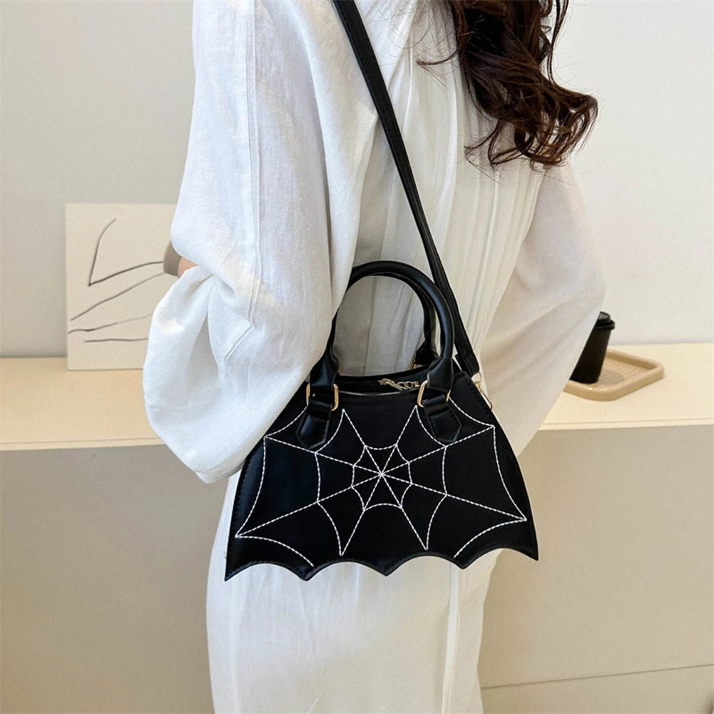 Black bat-shaped handbag with spider web design held by a person wearing a white outfit.