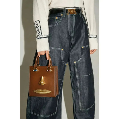 A person wearing dark blue wide-leg denim jeans with contrast white stitching, a white knit sweater, and holding a brown leather handbag with gold-tone hardware and a nose-shaped charm.