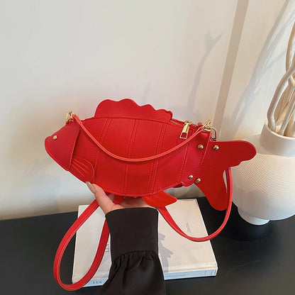 A bright red fish-shaped shoulder bag with gold-tone hardware, held by a hand over a white book on a dark table, with a white vase in the background.