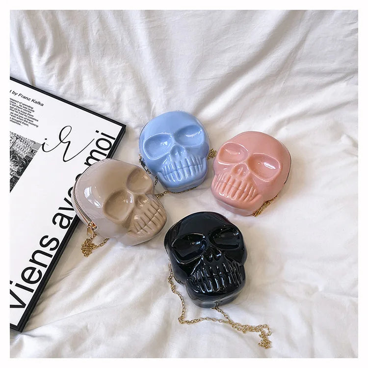 Four skull-shaped crossbody bags in beige, light blue, pink, and black, arranged on a white fabric background next to a magazine.