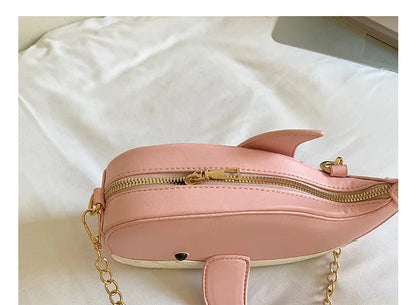 A pink whale-shaped crossbody bag with gold-tone hardware and a gold chain strap, resting on a white fabric background. A text overlay at the bottom reads 'THE DETAILS SHOW'.